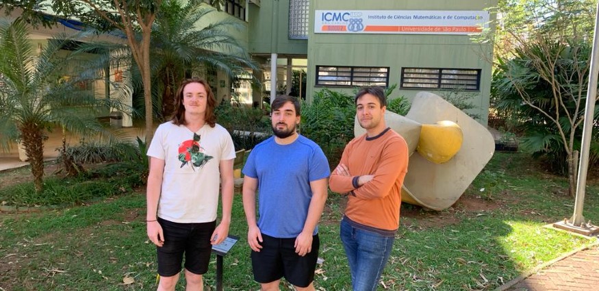 Three white males stand in front of trees and a building.